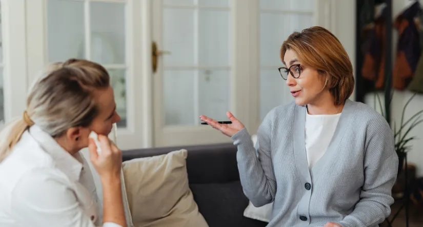 Two mature women talking on a cozy gray sofa in a bright living room; one woman with blonde ponytail wiping tears with tissue, the other with short red hair and glasses gesturing supportively with pen in hand, warm empathetic discussion atmosphere