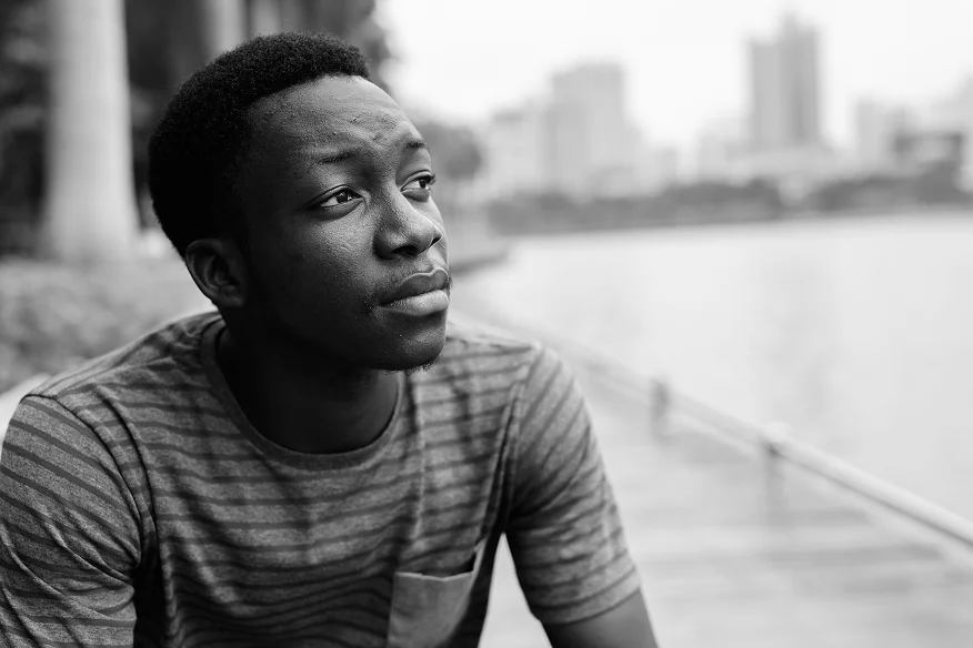 A young man in a striped shirt sitting outdoors by a lake, looking thoughtfully into the distance.