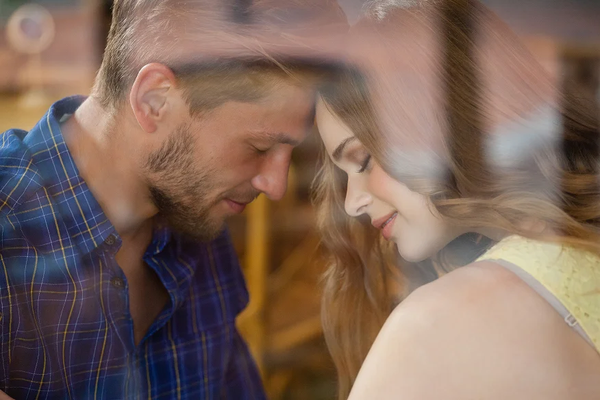 A young couple touching foreheads with eyes closed, showing an intimate moment of connection through a glass window reflection.