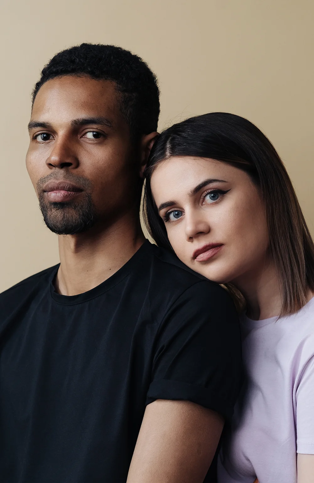 A young man in a black t-shirt and a woman in a lilac top leaning her head on his shoulder against a tan background